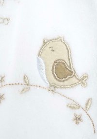 Búho bordado con detalles en beige y blanco, situado sobre un fondo de tela blanca, acompañado de sutiles acentos florales en colores claros.