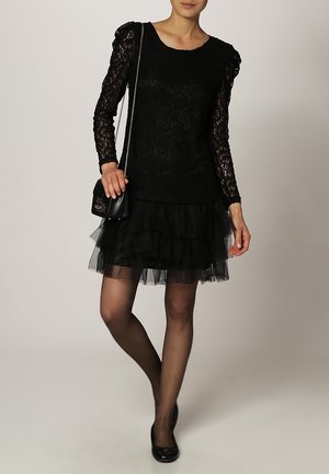 Zwarte kanten top met lange mouwen en textuurdetails, gelaagde zwarte tule rok, zwarte maillot en platte schoenen. Kleine zwarte schoudertas.