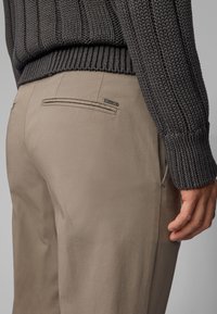 Pantalon beige à texture lisse, doté de poches arrière et de subtils accents métalliques, assorti à un pull en maille foncé au design texturé.