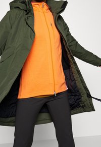 Giacca impermeabile verde sopra una felpa arancione con zip e tasche, abbinata a leggings neri. La giacca ha un cappuccio e una fodera a contrasto.