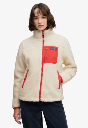 Joven mujer con chaqueta de forro polar crema con cremallera roja y detalles en los bolsillos, de pie con una mano en el bolsillo contra fondo blanco.