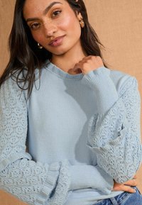 Svetlo modra pulover s enotno telo in pufastimi rokavi z čipkastim vzorcem; reberasti patent in ovratnik dodajata teksturo in podrobnosti.