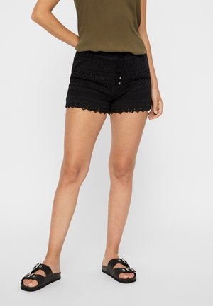 Person iført sorte hæklede shorts, olivengrøn ærmeløs top og sorte sandaler med dobbelte sølvspænder, stående med den ene hånd på hoften.