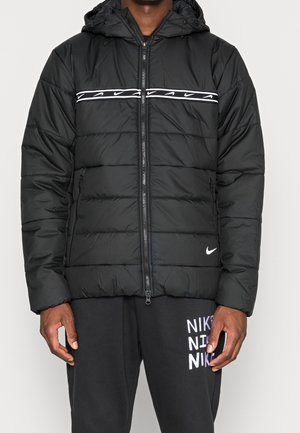 Mężczyzna ubrany w czarną, pikowaną kurtkę Nike z poziomym paskiem z logo Swoosh na klatce piersiowej oraz czarne spodnie dresowe Nike z napisem logo.