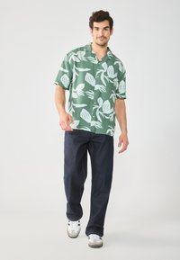 Kortermet grønn skjorte med knapper og hvitt bladmønster, kombinert med mørkeblå jeans og grå joggesko. Enkel, uformell stil.