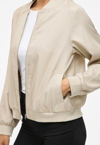 Beige bomberjakke laget av glatt stoff, med glidelås foran, ribbestrikkede mansjetter, to sidelommer og en avslappet passform.