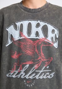 Nike sweatshirt i falmet sort med et stort gråt "NIKE" logo og en rød vingerpferd grafik, med rund hals og blød tekstur.