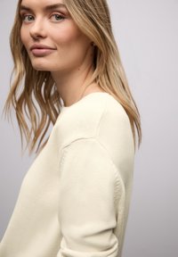 Pull tricoté beige clair avec un col rond et des détails de coutures subtils, porté par un mannequin aux cheveux ondulés. Texture lisse, design minimaliste.