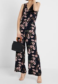 Combinaison fleurie en tissu bleu marine à motifs floraux roses. Elle présente un décolleté en V, des jambes larges, et un sac à main noir avec une poignée orange.