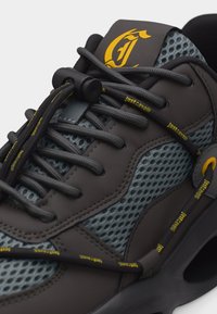 Μαύρο και γκρι αθλητικό παπούτσι με ύφασμα από δίχτυ, κίτρινο κείμενο «Just Cavalli» στα κορδόνια, κλείδωμα κορδονιών με σούστα και κίτρινο στιλιζαρισμένο λογότυπο στη γλώσσα.