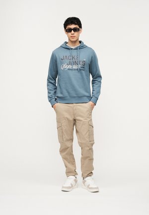 Ung man med svarta solglasögon, blå grafisk hoodie, beige cargobyxor och vita sneakers står med händerna i fickorna mot en enfärgad bakgrund.