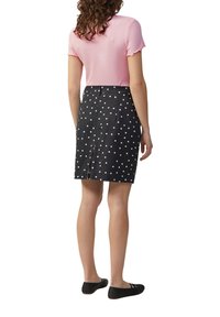 Frau in einem rosa Kurzarmoberteil, schwarzem knielangem Rock mit Punkten und Schlitz hinten, und schwarzen flachen Schuhen, die mit dem Rücken zur Kamera steht.