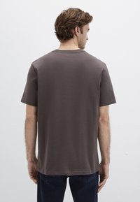 Braunes Baumwoll-T-Shirt mit kurzen Ärmeln, einem Rundhalsausschnitt und geradem Schnitt. Sichtbare Nahtdetails an Schulter und Rücken. Getragen mit dunklen Denim-Jeans.