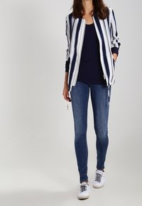 Chaqueta bomber a rayas en azul marino y blanco, combinada con una camiseta azul marino y jeans ajustados azules; zapatillas blancas con cordones negros, de textura suave.