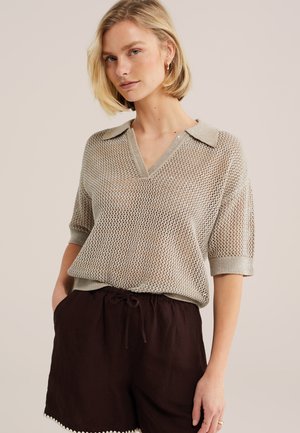 Blondínka v béžovom pletenom tričku s krátkym rukávom a golierom a v tmavohnedých šortkách so šnúrkou stojaca s rukami v kapsách.