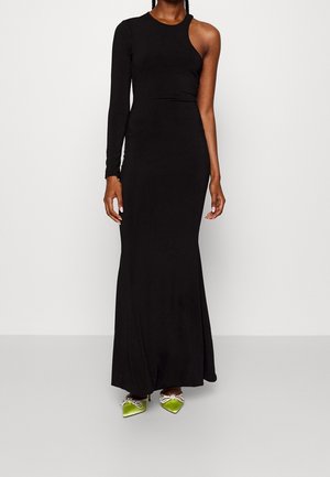 Frau trägt ein langes, schwarzes Kleid mit einem Ärmel und grüne spitze Schuhe mit dekorativen Schleifen, steht vor weißem Hintergrund.