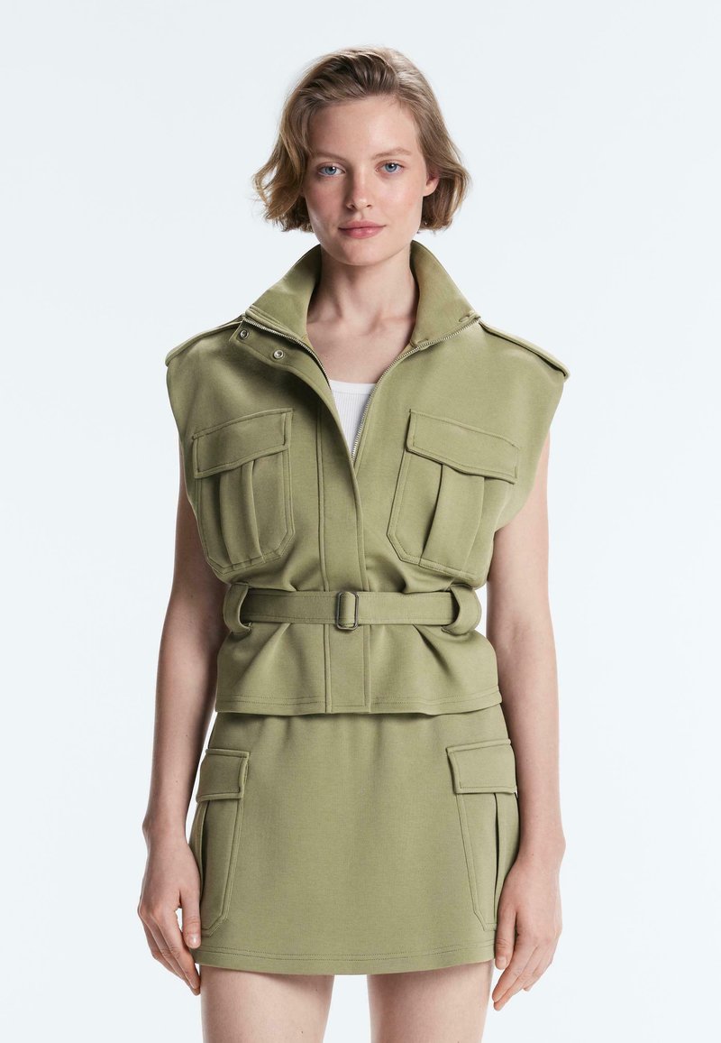 Femme aux cheveux blonds courts portant une veste sans manches kaki ceinturée avec de grandes poches et une mini-jupe assortie avec des poches latérales.