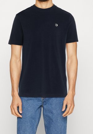 Mann in einem schlichten schwarzen T-Shirt mit kurzen Ärmeln und blauen Denim-Jeans, der mit entspannten Armen an den Seiten steht.