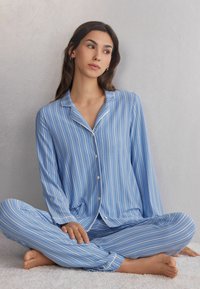 Lichtblauw pyjama set met witte strepen, bestaande uit een button-up top met een kraag en lange mouwen, gecombineerd met bijpassende broek. Zachte stoftextuur.