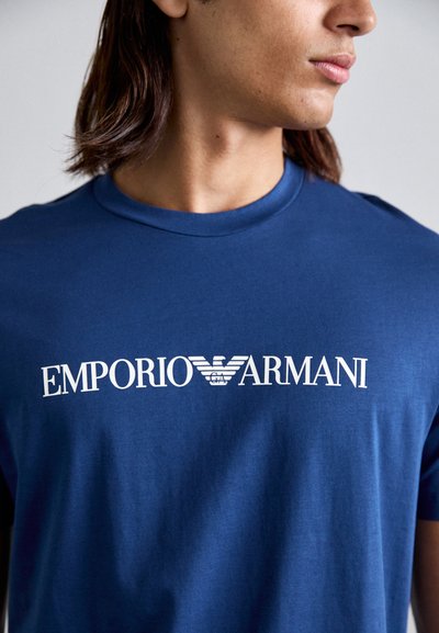 Modra bombažna majica s kratkimi rokavi in krožnim izrezom. Ima bel logotip "EMPORIO ARMANI" čez prsi z značilnim dizajnom orla.