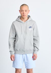 Siva zip-up kapuca z kengurujem žepom, s plavim Nike logotipom i natpisom. Uparen s svijetloplavim šortsem, izrađenim od mekog materijala.