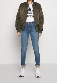 Oliwkowa bomberka, biała koszulka z czarnym logo Adidasa, dżinsy oraz białe sneakersy z niebieskimi i zielonymi akcentami.