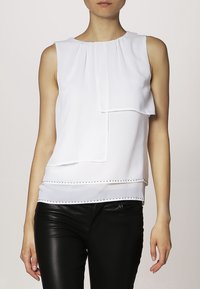 Blusa bianca senza maniche con design a strati, pieghe al collo e una linea decorativa di borchie metalliche lungo l'orlo. Tessuto di consistenza liscia.
