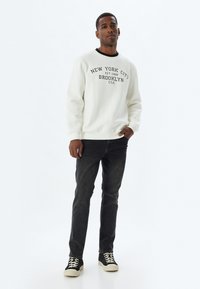 Sudadera blanca con texto negro, cuello acanalado negro, combinada con jeans oscuros y zapatos negros con suelas blancas. Look simple y casual.