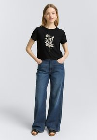 Svart t-shirt med ett ljust blommönster, kombinerad med vida blå jeans. Klädseln har en avslappnad passform och en avslappnad stil.