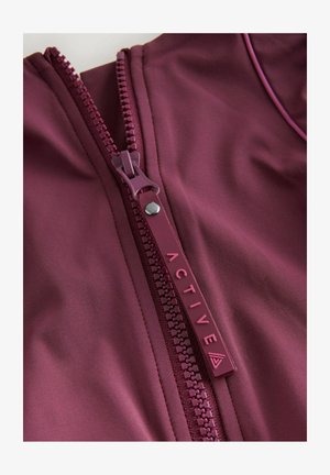 Close-up van een gedeeltelijk open ritssluiting van een bordeauxrode jas met een bijpassend ritskoordje waarop "ACTIVE" is gegraveerd en een klein driehoekig logo.