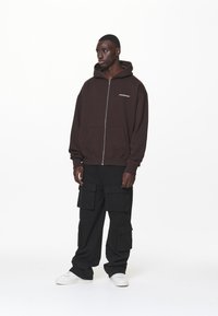Felpa con zip marrone, con tasca frontale e logo bianco, abbinata a pantaloni cargo neri con più tasche e sneaker bianchi.