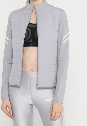 Femme portant une veste matelassée gris clair avec des bandes réfléchissantes sur les manches, un soutien-gorge de sport noir et des leggings Nike gris clair.