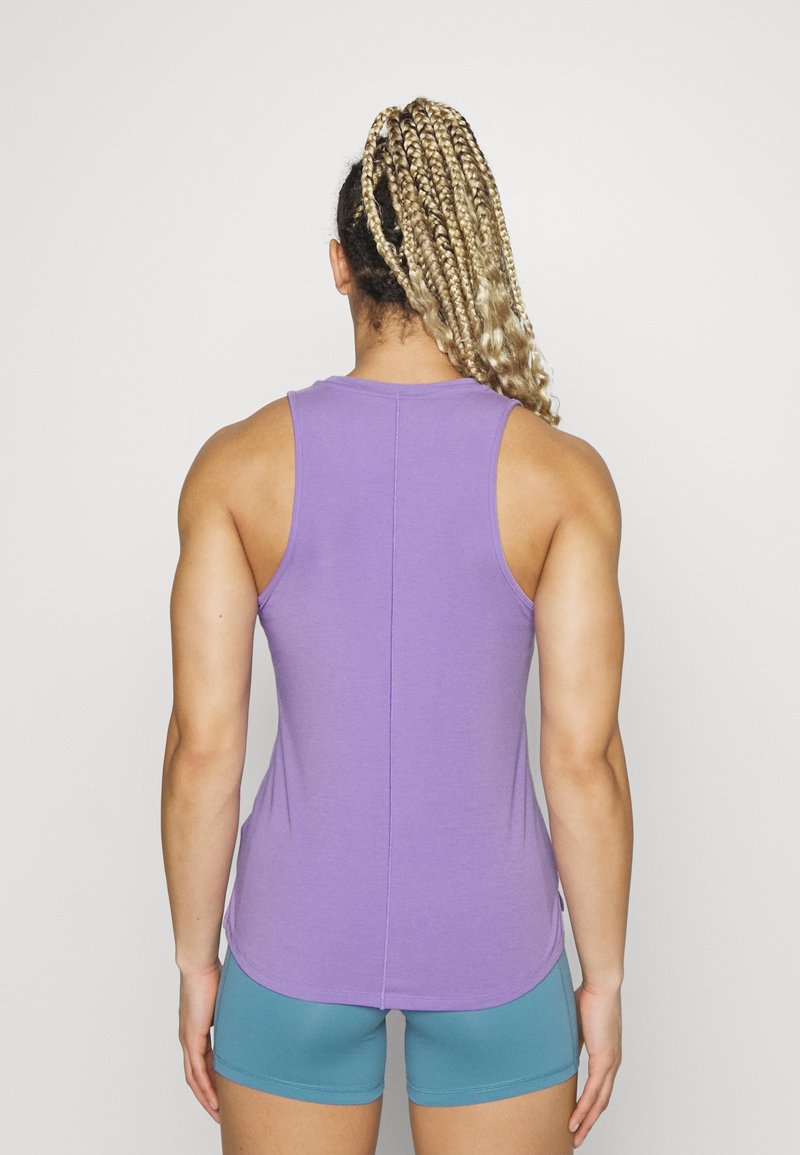 Personne athlétique aux cheveux tressés portant un haut sans manches violet et un short bleu sarcelle, debout de dos sur un fond uni.