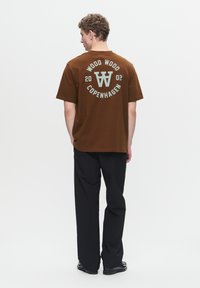 Brown cotton t-shirt featuring a circular graphic with "WOOD WOOD COPENHAGEN 2002" in light blue on the back. Black trousers complete the look.