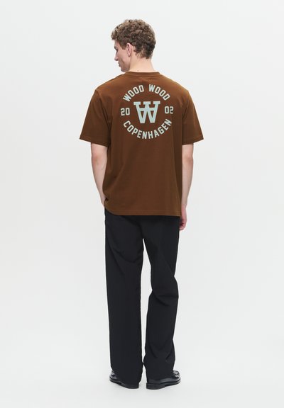 T-shirt in cotone marrone con una grafica circolare con "WOOD WOOD COPENHAGEN 2002" in blu chiaro sulla schiena. Pantaloni neri completano il look.