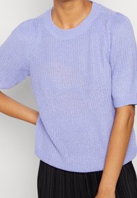 Person in einem helllila gestrickten kurzärmligen Pullover mit Rundhalsausschnitt und einem schwarzen Faltenrock.