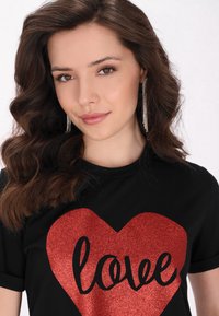Sort t-shirt med et stort rødt glitrende hjerte, hvor ordet "love" står i sort kursiv. Krøllet hår og lange øreringe fuldender looket.