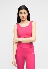 Γυναίκα στέκεται με σταυρωμένα τα χέρια, φορώντας φωτεινή ροζ αθλητική κοντή μπλούζα και αντίστοιχα ψηλόμεσα ποδήλατικά σορτς με λογότυπα EA7.