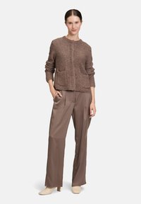 Frau steht mit der Hand in der Tasche, trägt einen braunen strukturierten Pullover, lockere braune Satin-Hosen und beige, bezogene Stiefeletten mit Absatz.