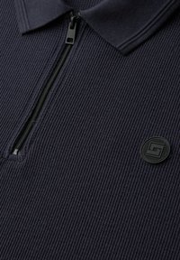 Nærbillede af mørkt ribstrikket stof med en sort lynlås og et rundt, tekstureret logopatch nær kraven.