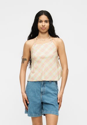 Femme aux longs cheveux foncés portant un haut sans manches à carreaux pastel et un short en jean bleu taille haute, debout devant un fond uni.