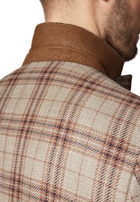 Brązowa kratkowana kurtka z teksturowanej beżowej tkaniny i kontrastującym brązowym zamszowym kołnierzem. Widoczne szwy wzdłuż kołnierza.