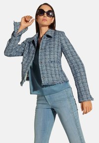 Lichtblauwe tweed jas met franje, voorzien van zilveren knopen en voorzakken. Draagt over een blauwe top en gecombineerd met lichtblauwe jeans.