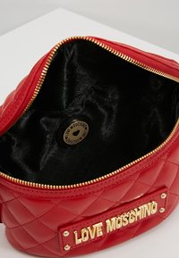 Červená prošívaná kabelka s zlatým zipem, značkovou destičkou na přední straně a černým interiérem s opakujícím se vzorem loga.