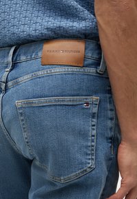 Close-up af en person iført blå jeans med en brun Tommy Hilfiger læderlap og et lille logo på baglommen.