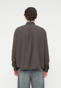 Σκούρο γκρι καρό που κάμισο με άνετη εφαρμογή, μανσέτες με κουμπιά και πλάτη με πιέτα. Φορεμένο με ξεθωριασμένο μπλε τζιν, που δείχνει από πίσω.