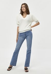 Weißer gestrickter Pullover mit V-Ausschnitt und puffierten Ärmeln, kombiniert mit hellblauen Schlagjeans und schwarzen High Heels. Insgesamt glatte Textur.