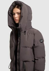 Padded bunda v tmavosivej farbe s veľkou, štruktúrovanou kapucňou. Obsahuje plný zips, šnúrky a značkovú nášivku na rukáve.