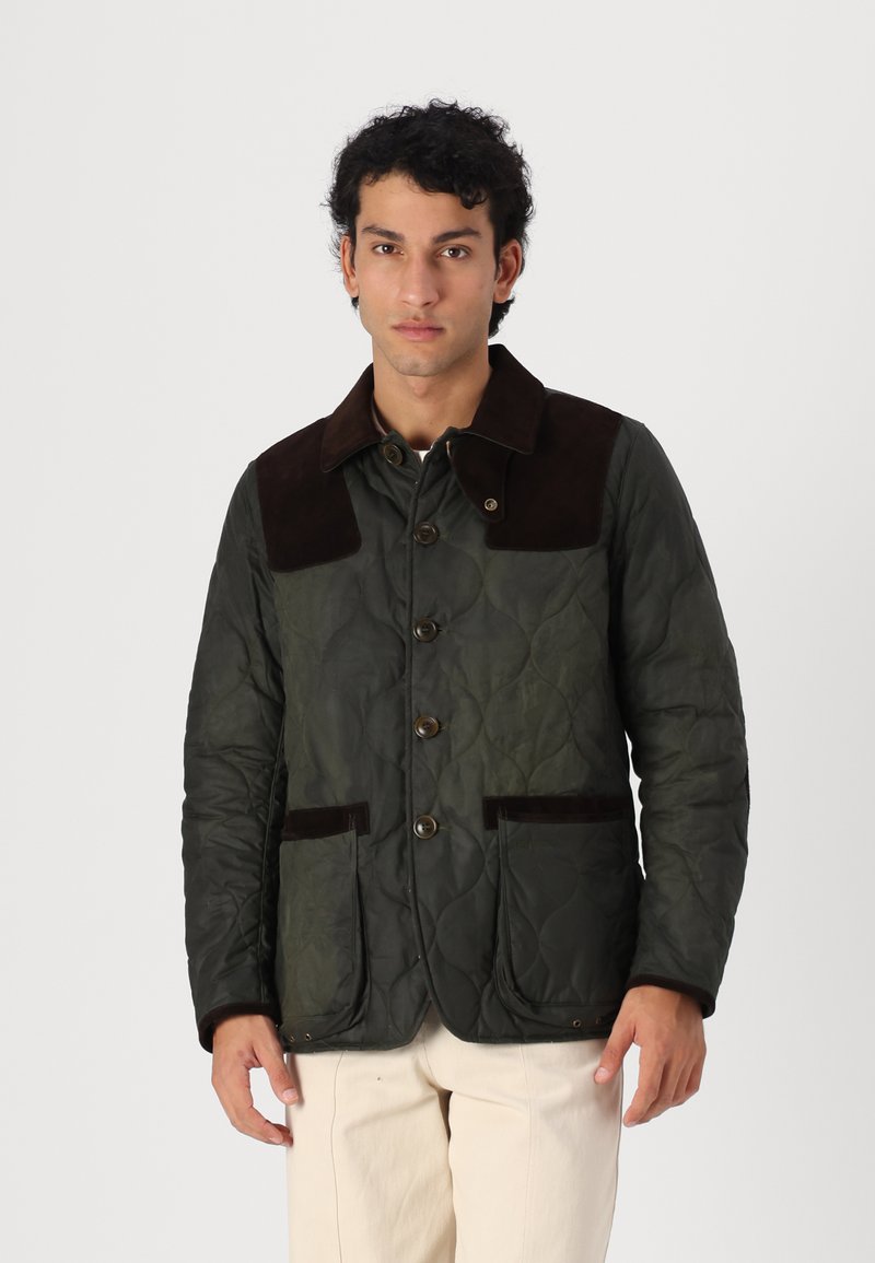 Ein Mann trägt eine dunkelgrüne Steppjacke mit braunem Kragen und Taschenverzierungen und steht vor einem einfachen hellen Hintergrund.