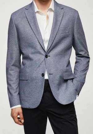 Mann trägt hellblauen, strukturierten Blazer über weißem Hemd und schwarzen Hosen, eine Hand in der Tasche, Oberkörper sichtbar, neutraler Hintergrund.